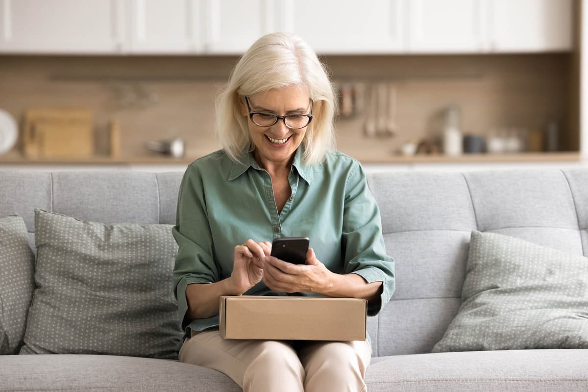 Smiling woman with glasses, sitting on a couch, using a smartphone with a cardboard box on her lap.