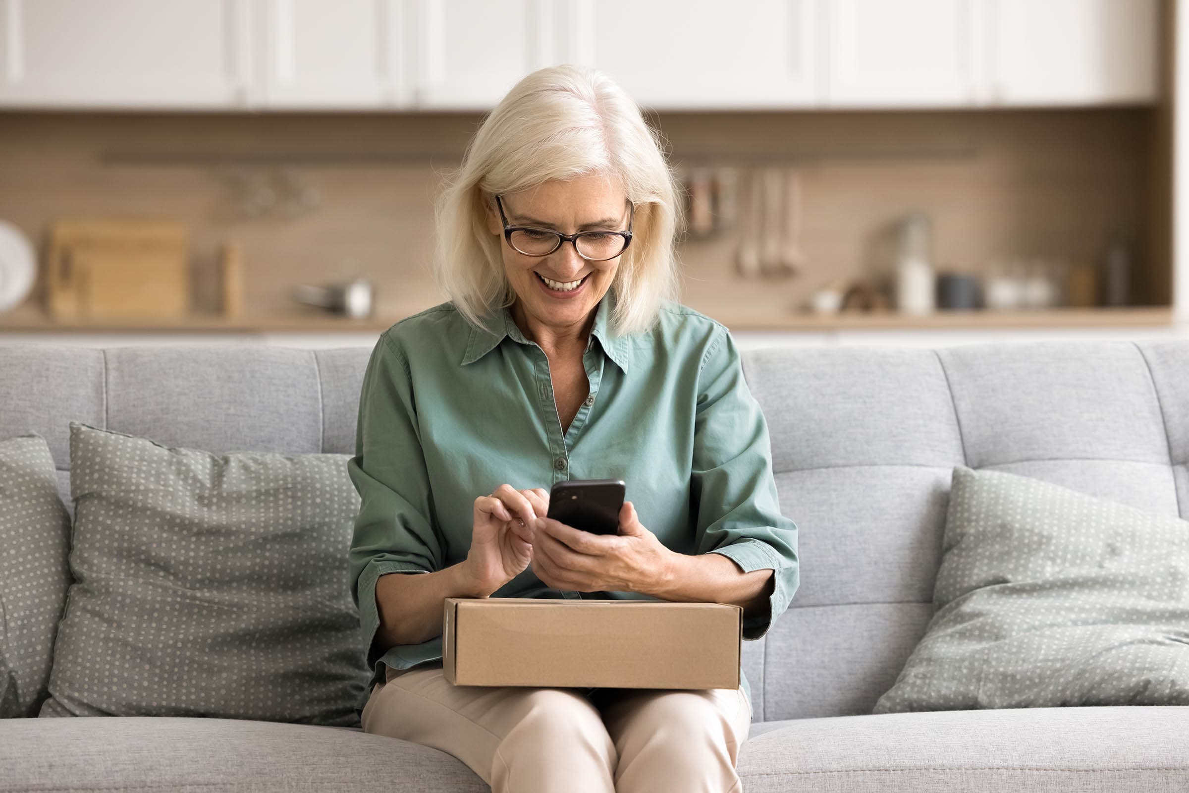 Smiling woman with glasses, sitting on a couch, using a smartphone with a cardboard box on her lap.