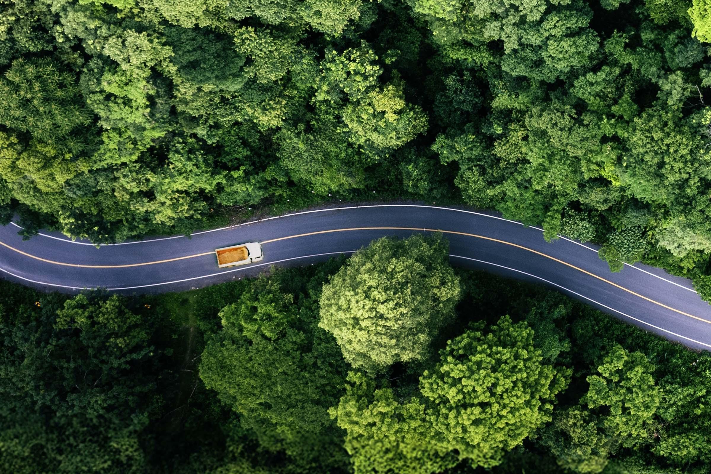 Aerial view of a winding road cutting through dense green forest with a single yellow truck driving along it.