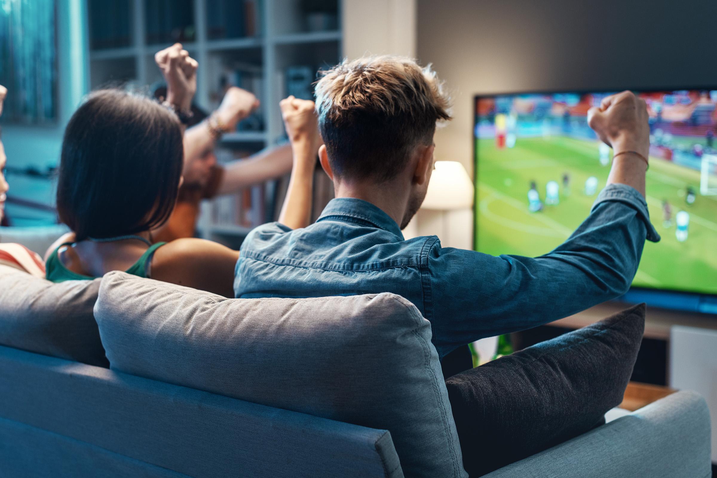 Friends on sofa cheer with raised arms while watching a soccer match on large TV in softly lit living room.