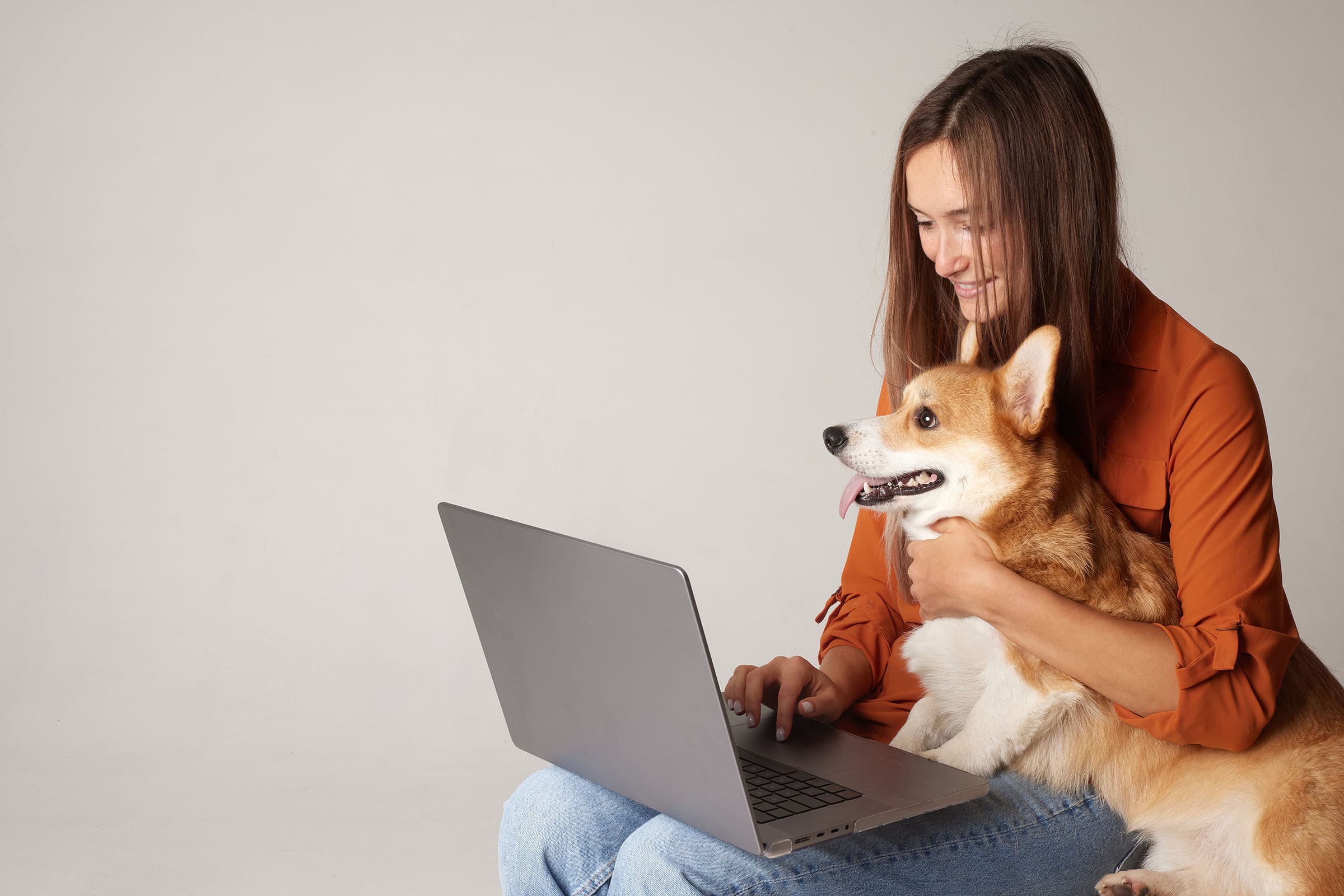 A person in an orange shirt sits beside a laptop, smiling and hugging a happy corgi dog.