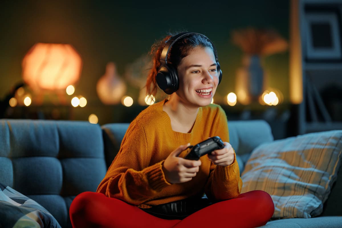 A person in a cozy room, wearing headphones and a sweater, is smiling while playing a video game with a controller.