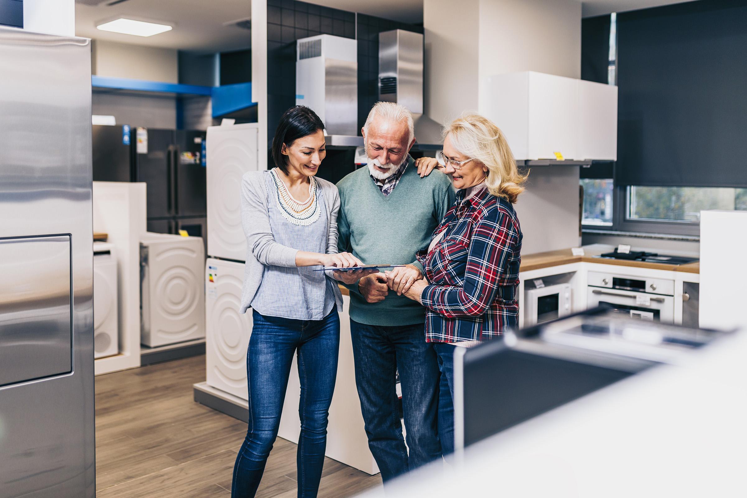 A female store employee shows product details on a tablet to an older couple in a modern appliance showroom.