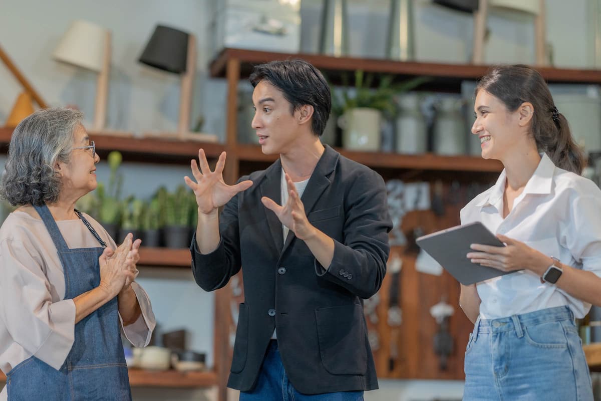 Three people engaged in a lively discussion. A woman in an apron, a man gesturing, and a woman with a tablet smile in a cozy setting.