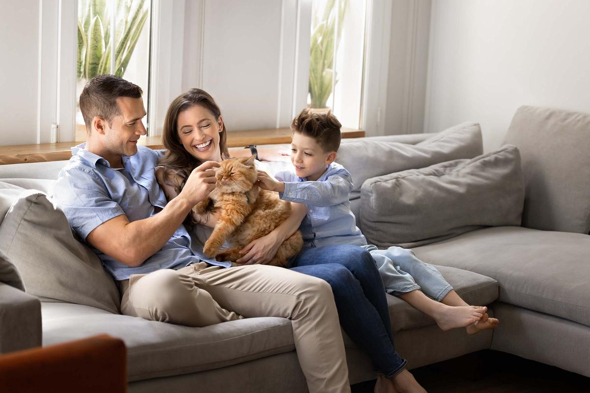 A smiling family of three sits on a couch petting a fluffy orange cat in a bright living room.