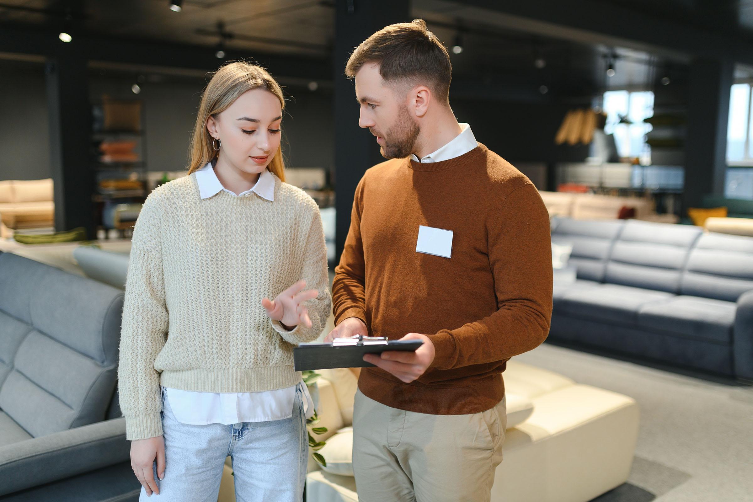 Woman talking to a male salesperson holding a clipboard in a furniture store with sofas in the background.