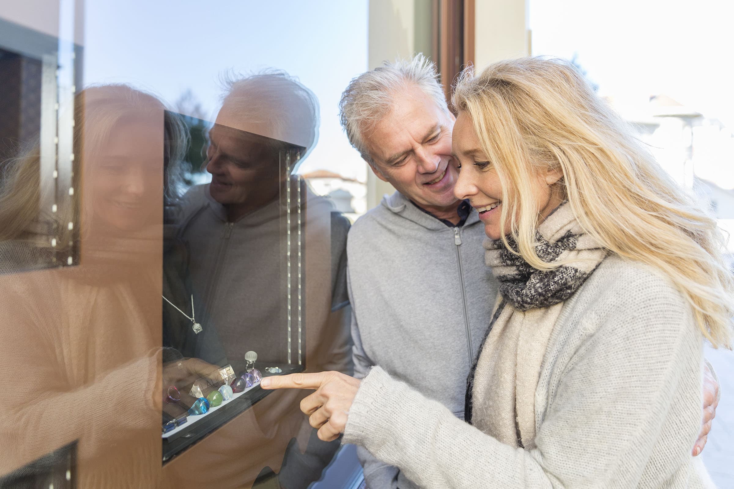 Smiling couple looking at jewelry through a store window, with the woman pointing at a display of colorful rings.
