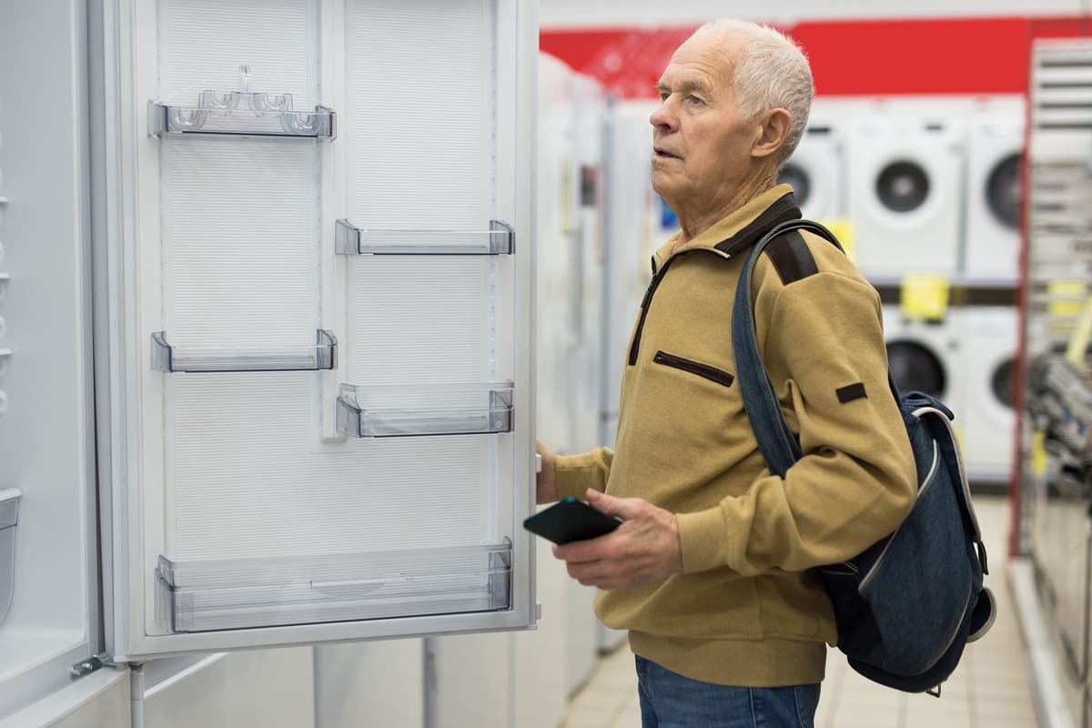 Older man holding a phone and inspecting the inside of an open refrigerator in an appliance store, wearing a backpack and beige jacket.