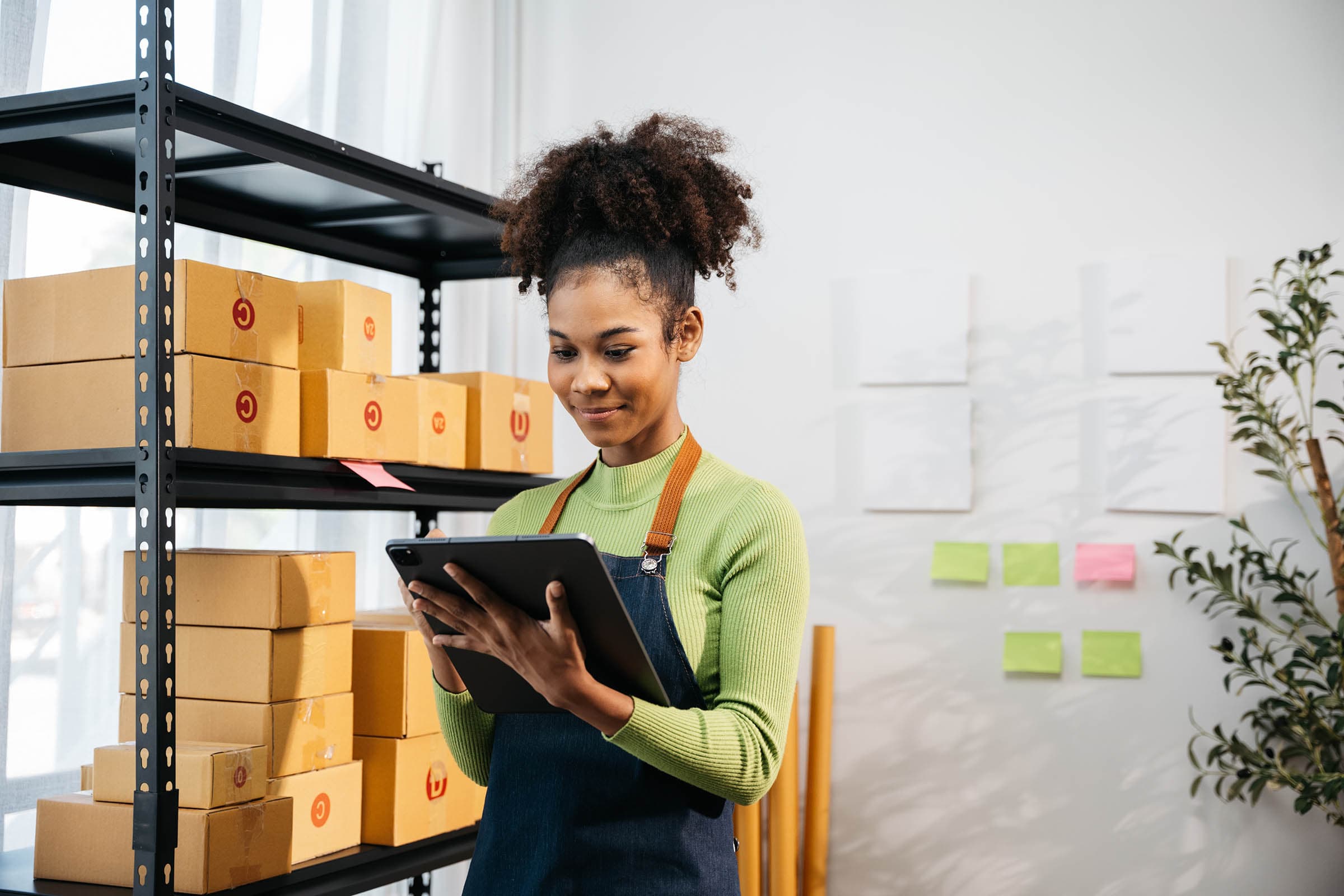 Woman with curly hair in a green sweater and apron uses a tablet in a room with shelves of boxes and sticky notes on the wall.