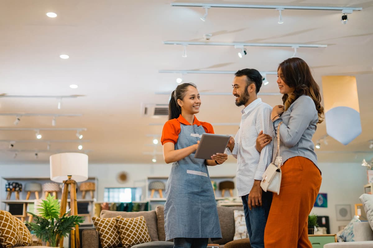 A couple in a furniture store talks with a smiling sales assistant holding a tablet, surrounded by various home decor items.