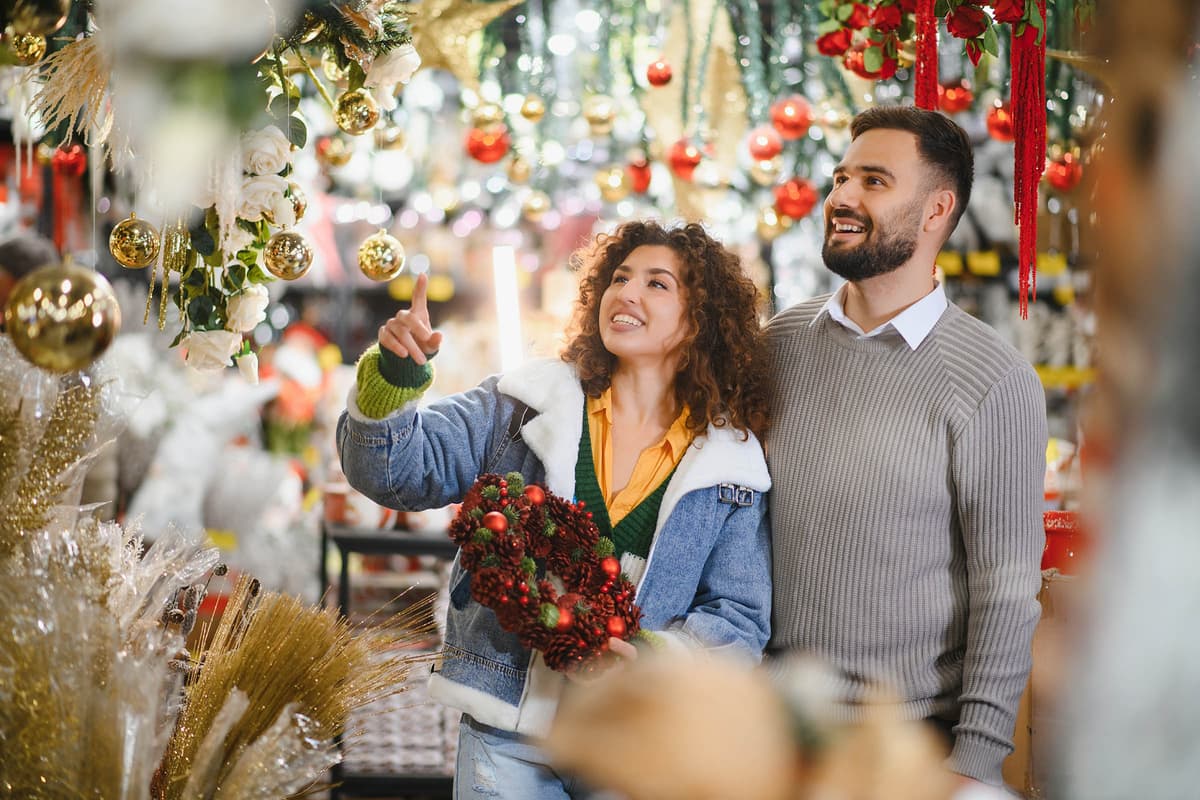 A joyful couple admires holiday decorations in a store, surrounded by festive ornaments and holding a decorative wreath.