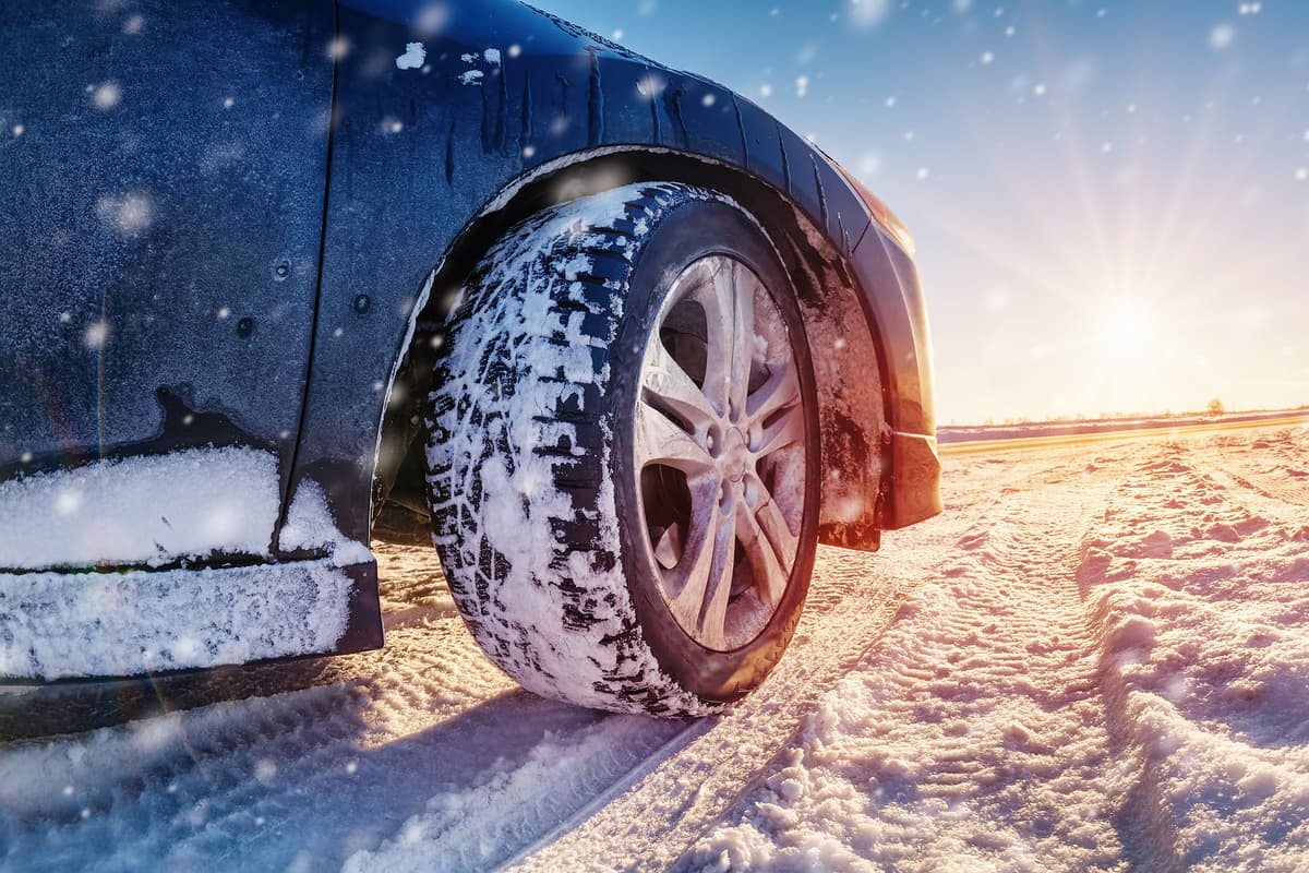 Close-up of a car tire covered in snow on a snowy road, with sunlight and falling snow in the background.