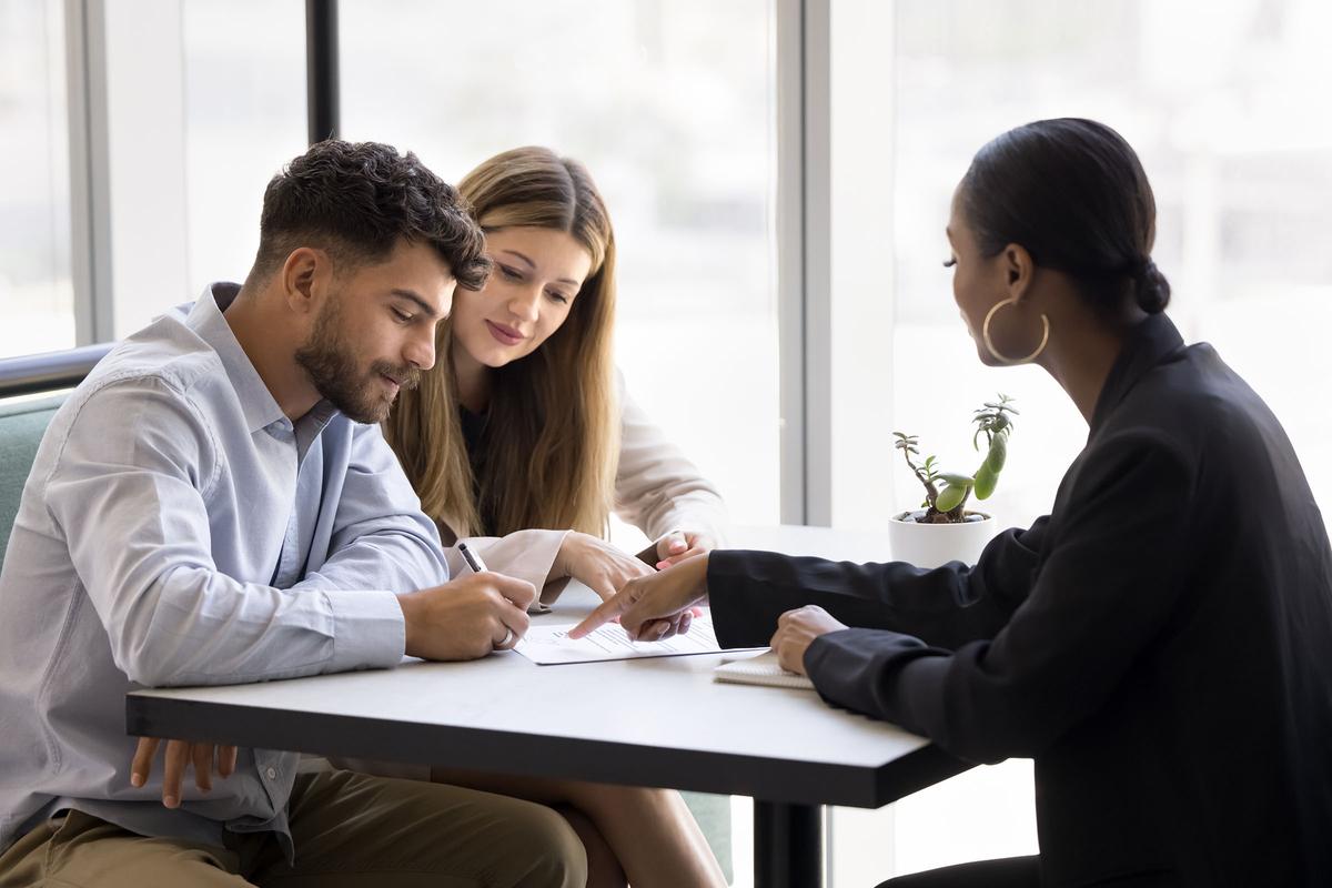 Couple signing a document at a table while a professional woman in a blazer points to the paperwork in a bright office setting.