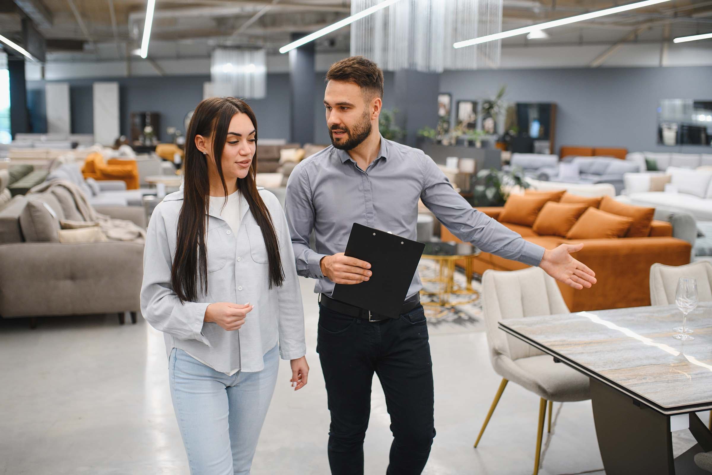 A woman and a man walk through a furniture store, with the man holding a clipboard and gesturing towards a display of tables and sofas.