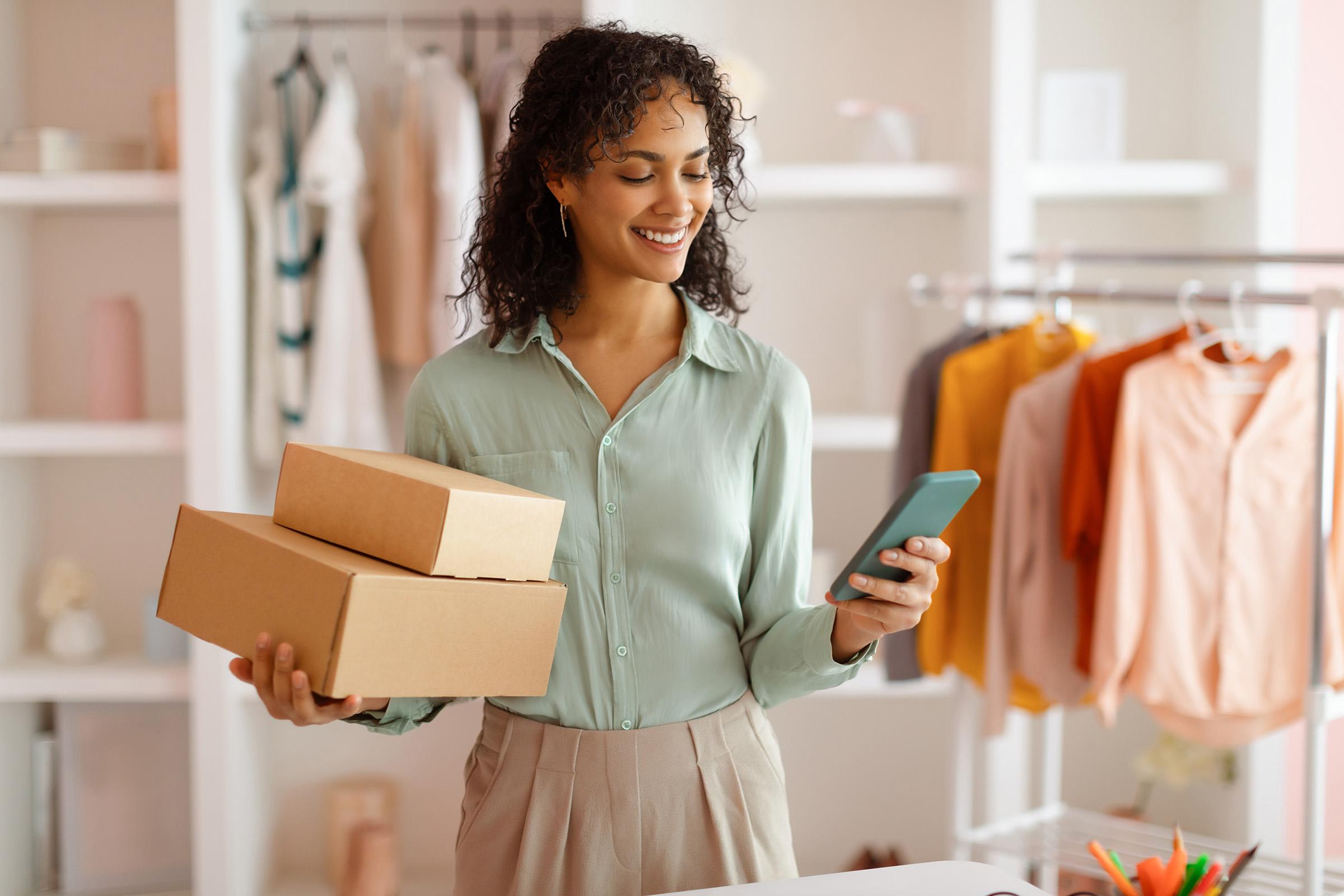 Smiling woman holding two cardboard boxes and a smartphone, standing in a clothing store or boutique.
