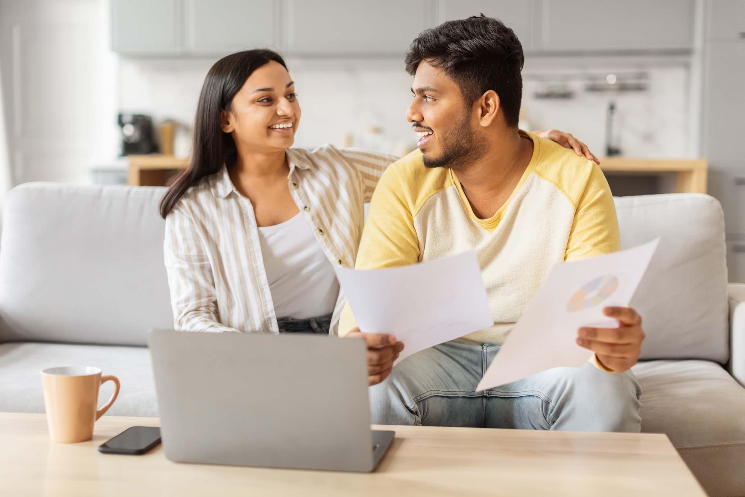 Smiling couple sitting on a couch with a laptop, holding documents and discussing something in a modern living room.