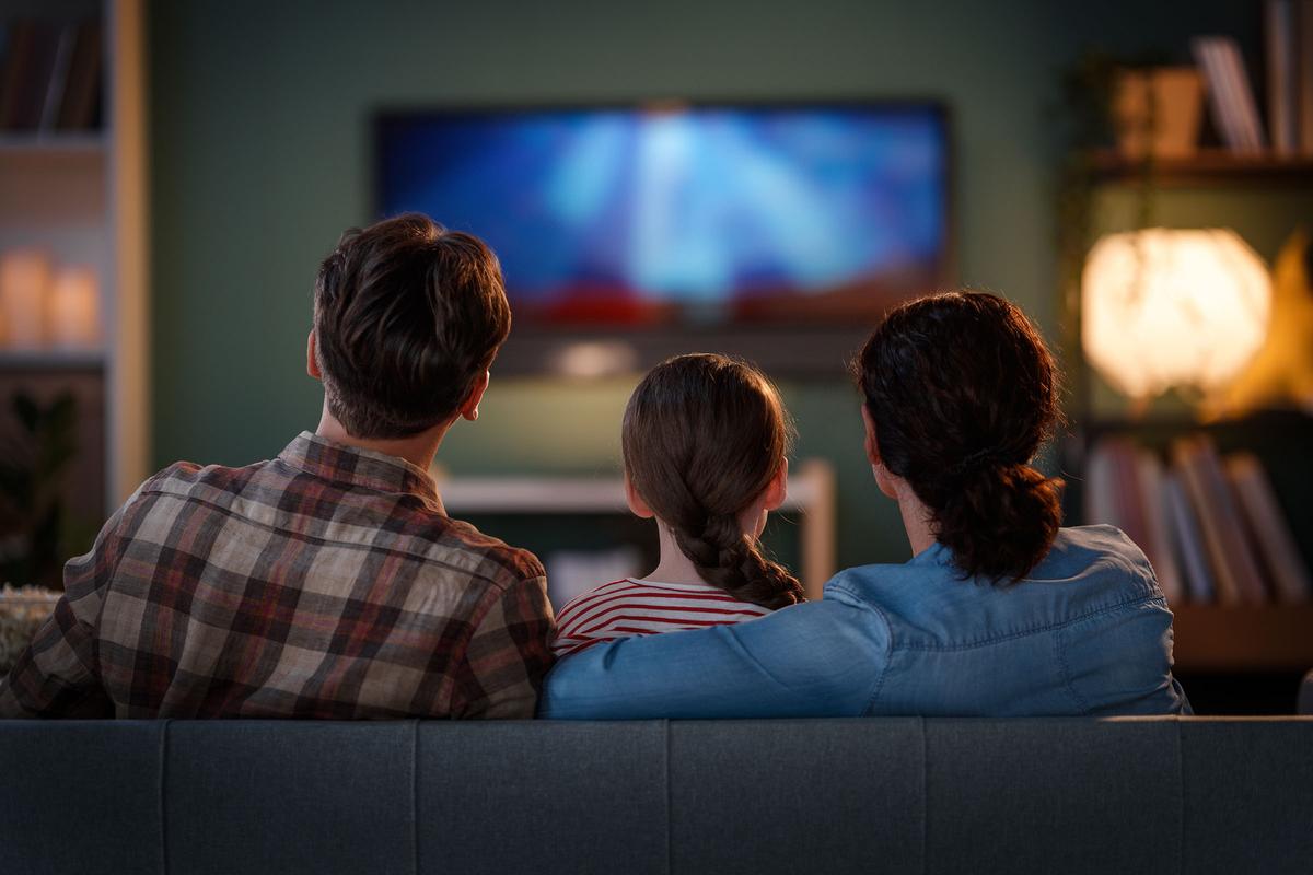 Three people sit on a couch with their backs to the camera, watching a television in a cozy, dimly lit living room.