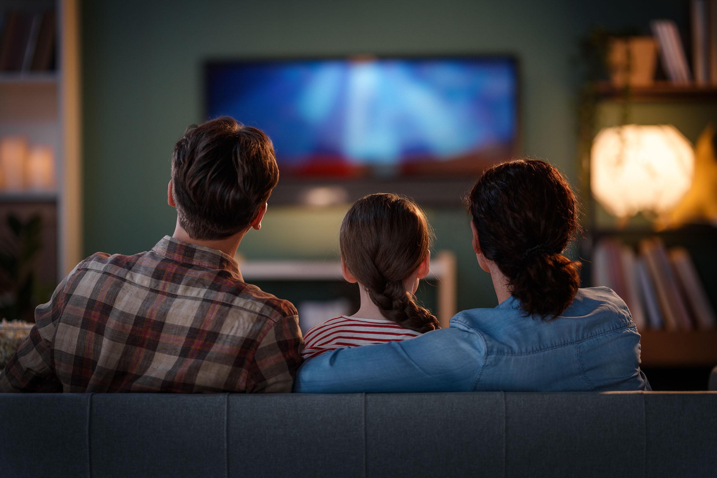 Three people sit on a couch with their backs to the camera, watching a television in a cozy, dimly lit living room.