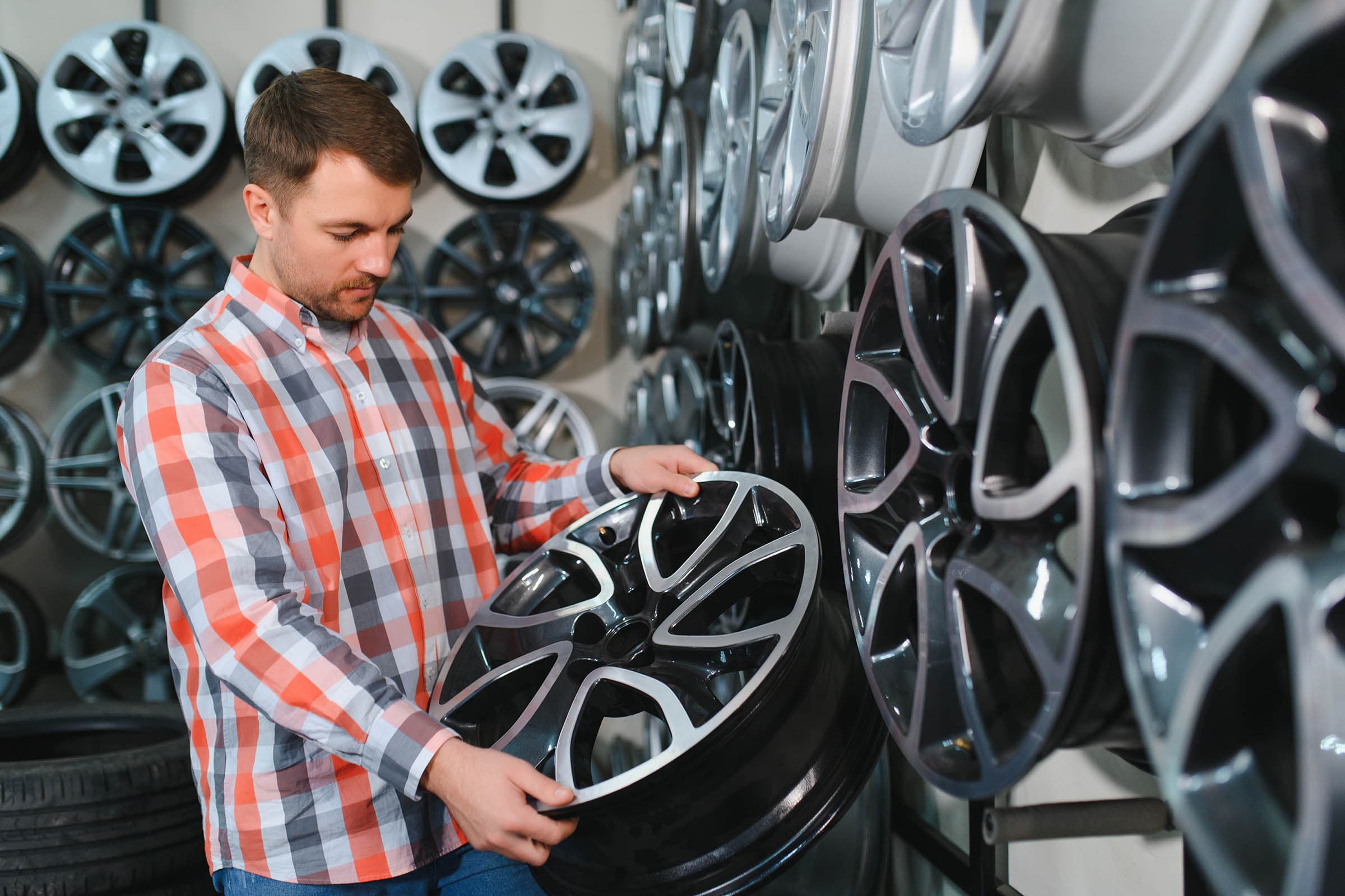 Man in plaid shirt examining a shiny car rim in a shop, surrounded by various wheel designs on display.