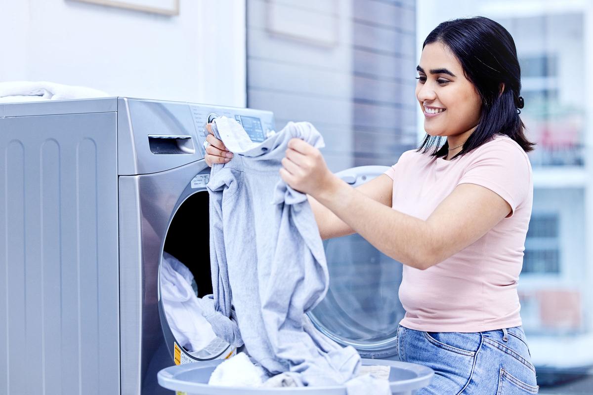 Smiling woman unloads a grey shirt from stainless front-load washer, placing laundry into a basket during household chores.