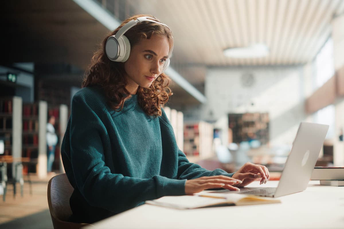 Woman in a sweater wearing headphones, focused on a laptop in a spacious library with bookshelves blurred in the background.