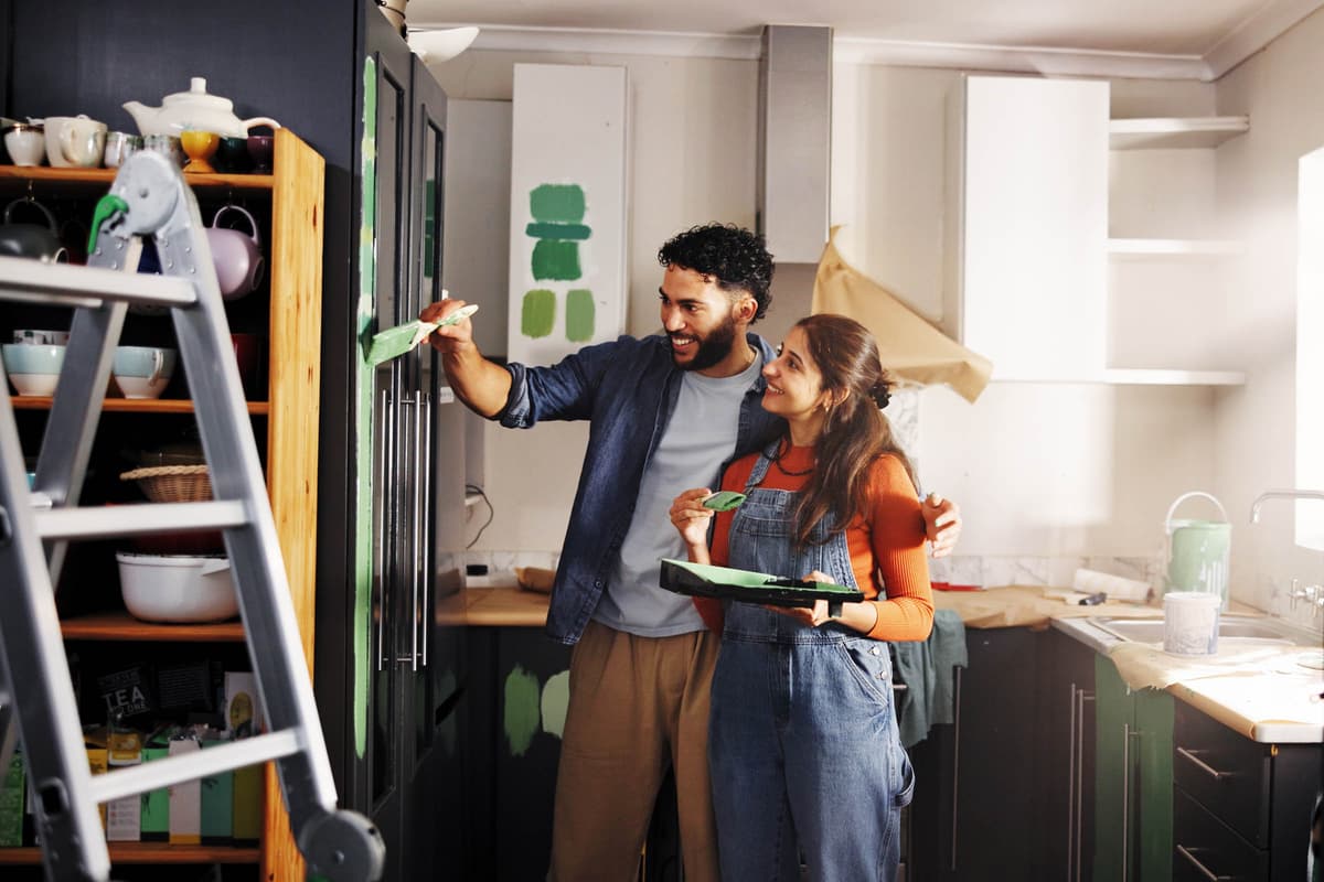 Couple painting a kitchen cabinet green, surrounded by painting supplies and a step ladder, smiling at their progress.