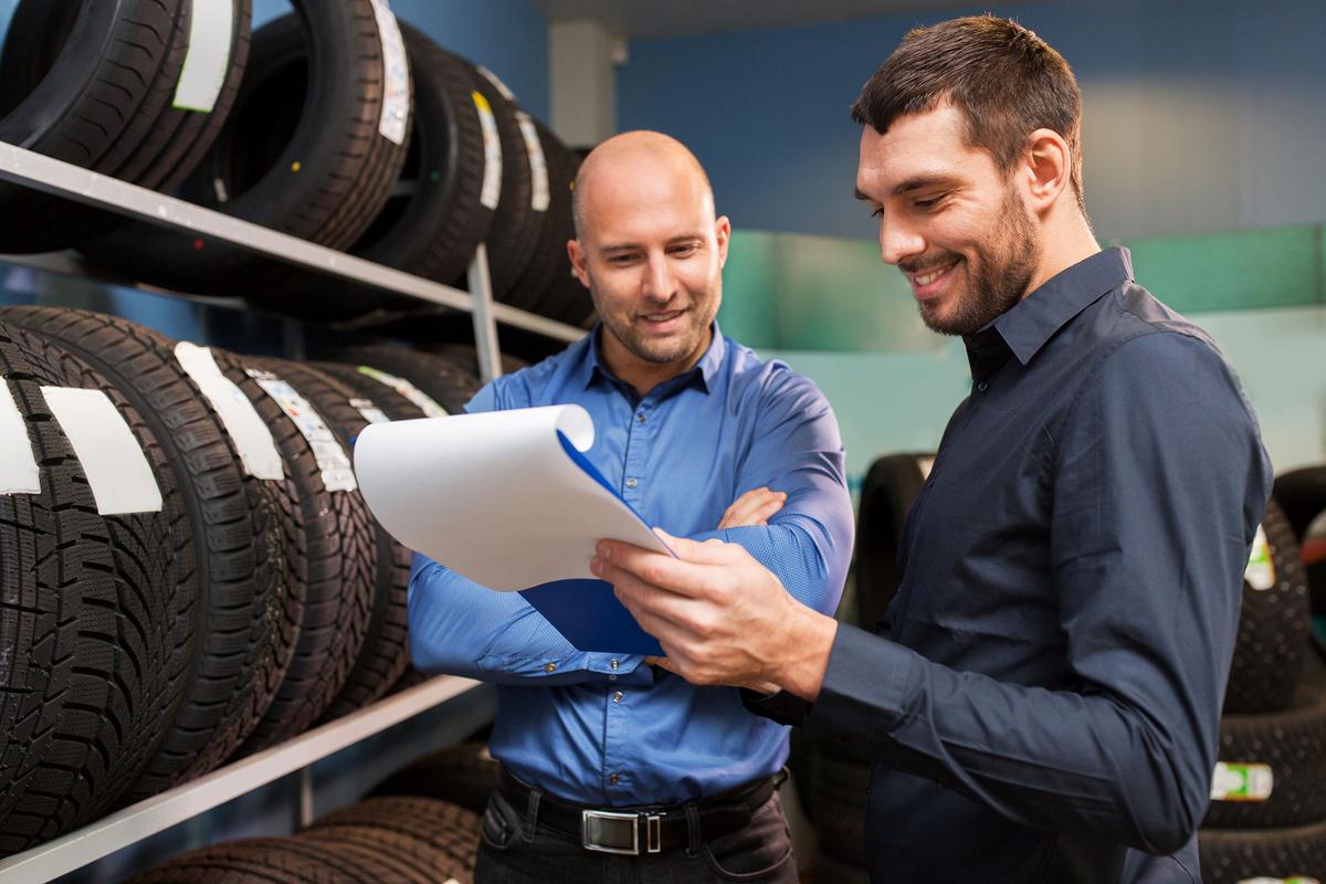 Two men standing in a tire shop, smiling while reviewing a document in front of shelves stacked with car tires.