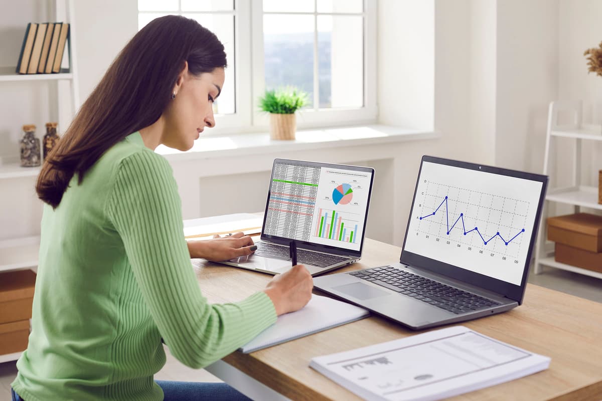 Woman in green sweater working at a desk with two laptops displaying charts and graphs, writing on a notepad.