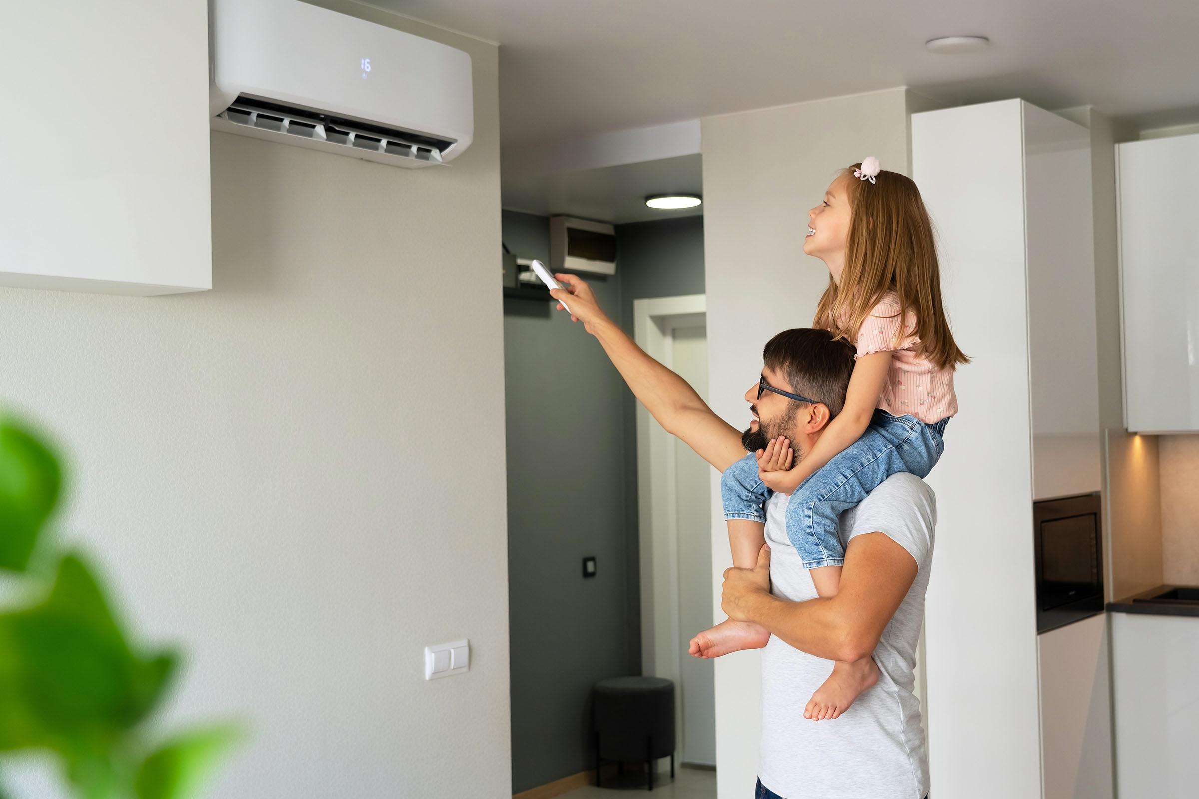 Man carrying a smiling girl on his shoulders as she points a remote at a wall-mounted air conditioner in a modern home.