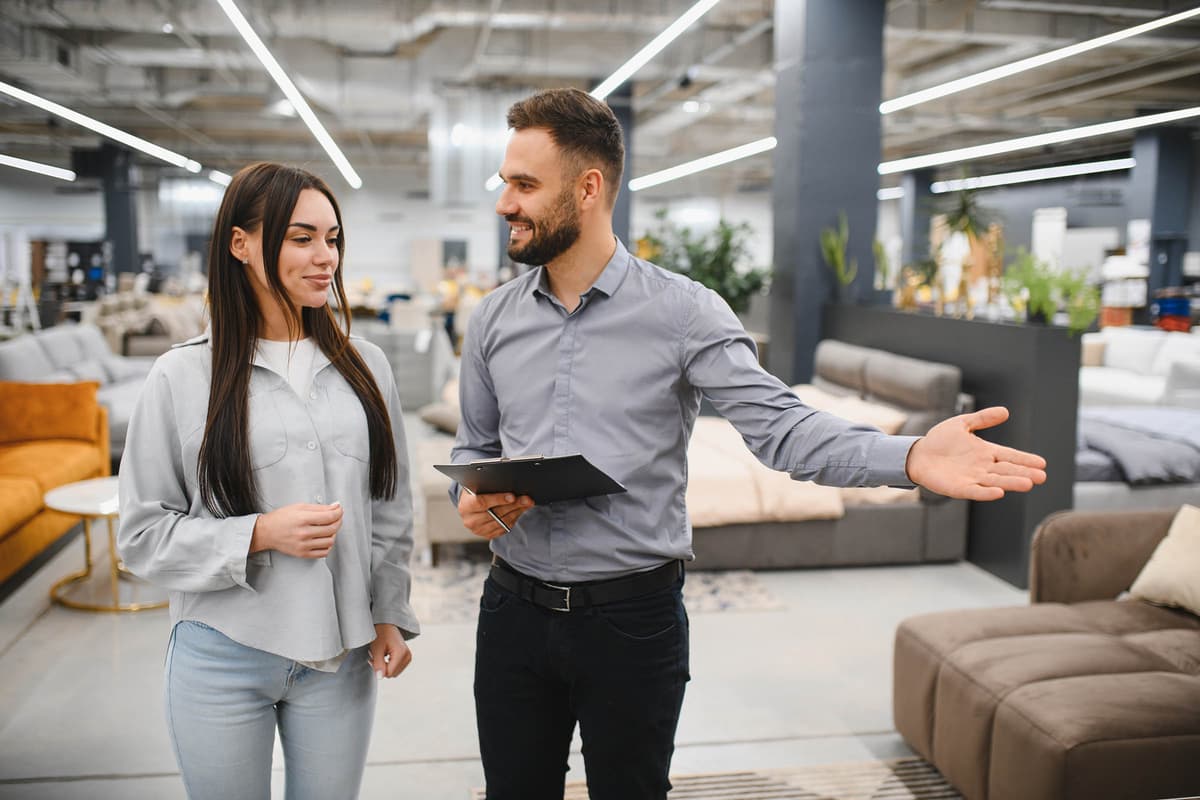 A man shows a woman around a furniture store, gesturing towards sofas and beds, while holding a clipboard.