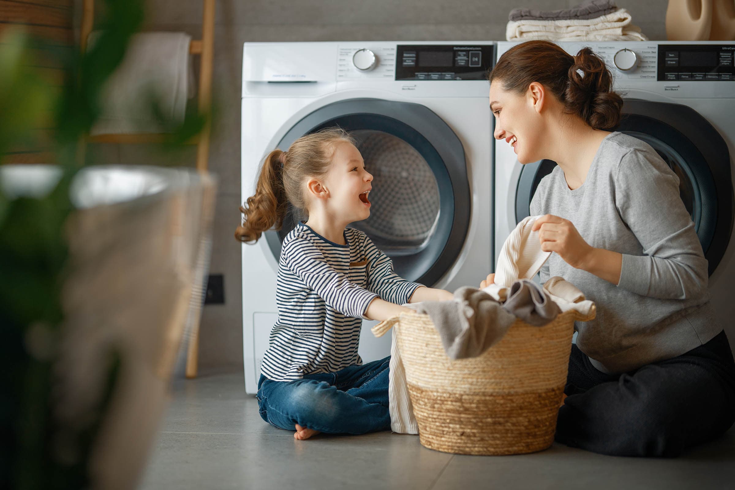 Mother and daughter smiling while doing laundry together in a modern laundry room with a basket of clothes and a washer and dryer.