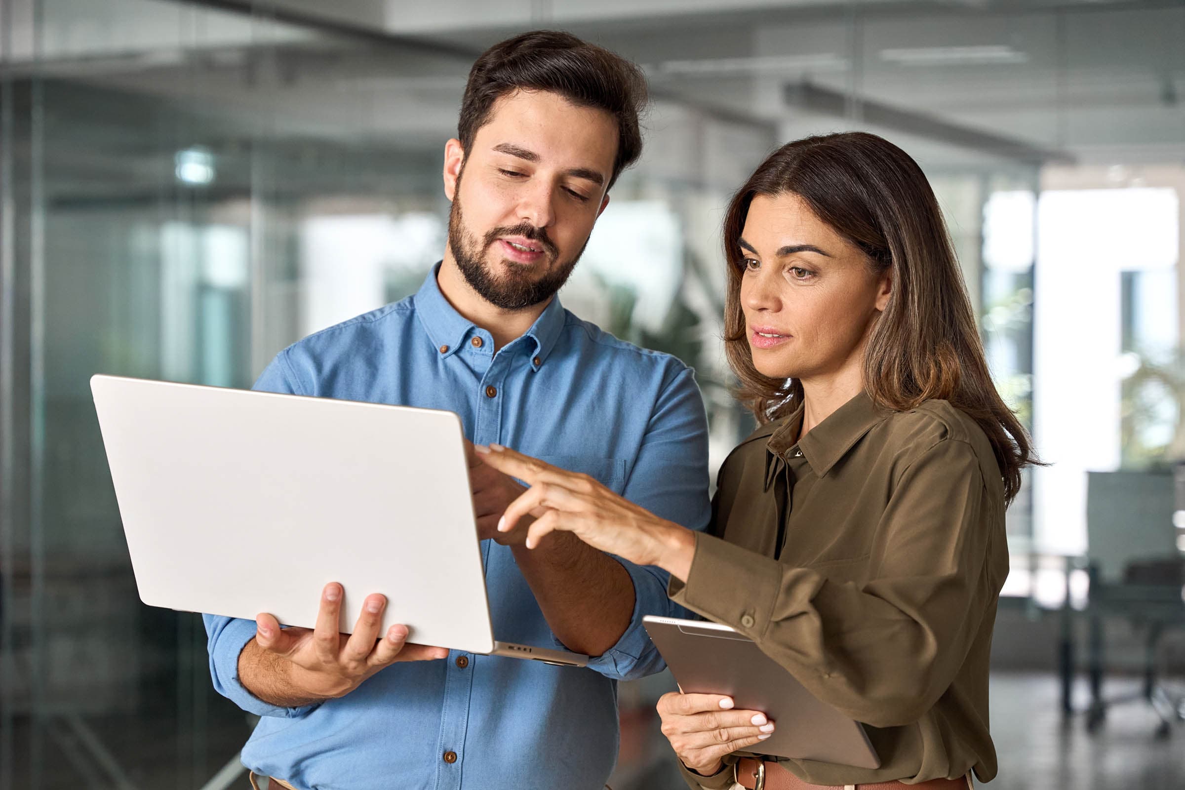 Two colleagues in a modern office discuss something on a laptop. One holds a tablet, while the other gestures at the laptop screen.