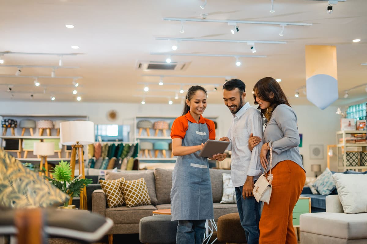 A store employee assists a smiling couple with a tablet in a furniture store, surrounded by sofas and shelves of products.