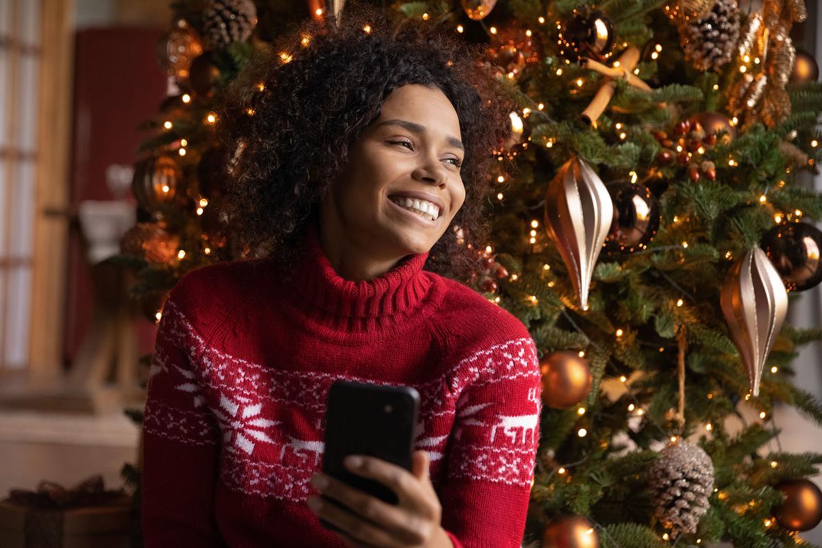 Smiling woman in red festive sweater holds smartphone beside Christmas tree glowing with lights and bronze ornaments.