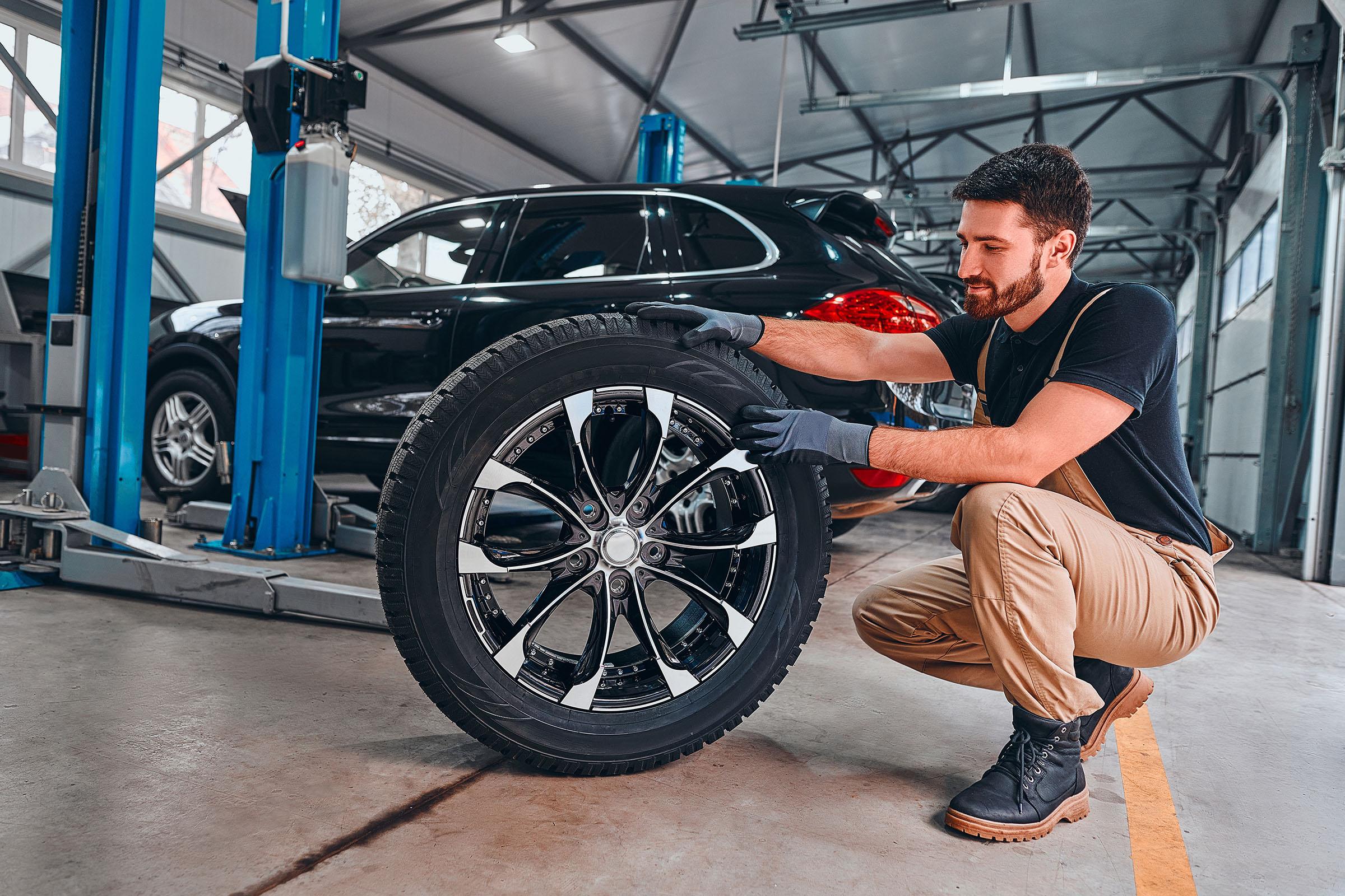 Mechanic in gloves kneels in auto shop, examining a large black tire with shiny alloy rim next to SUV on hydraulic lift.
