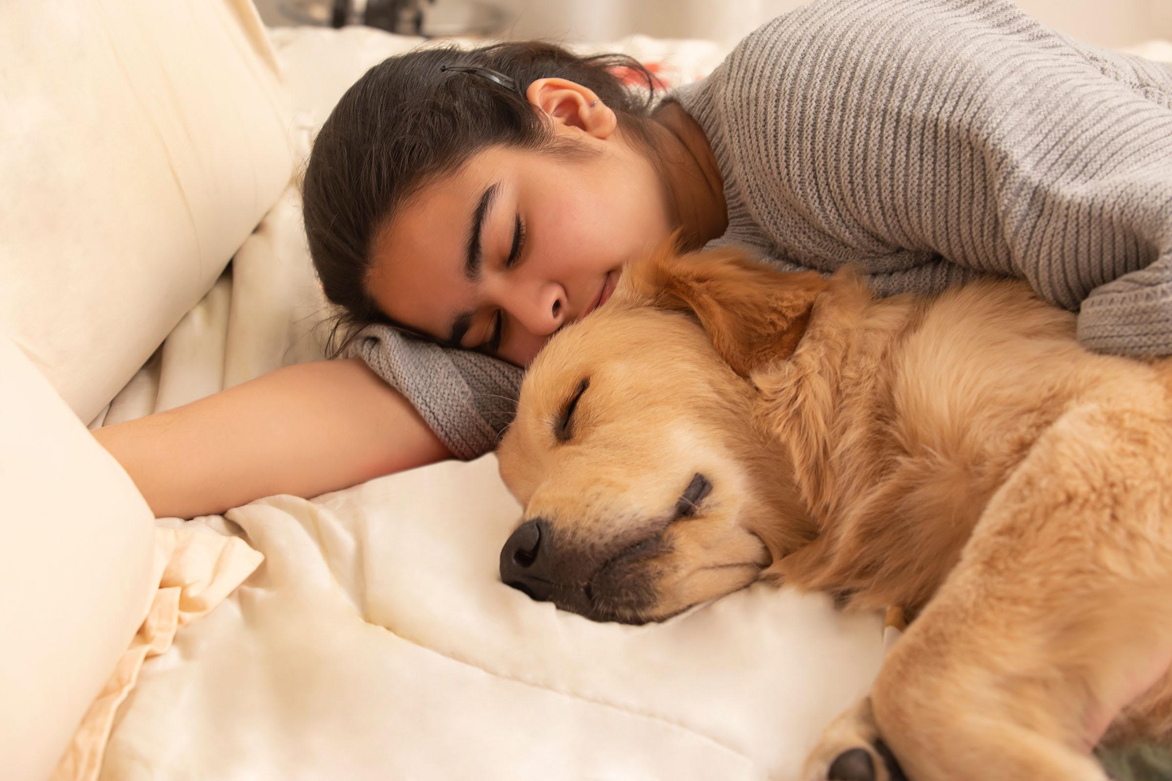 A woman lies on a bed cuddling a sleeping golden retriever, both appearing relaxed and peaceful.