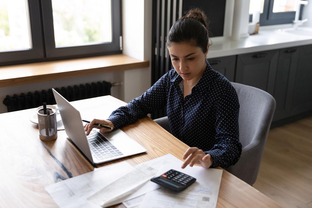 Woman in a dark polka dot blouse working at a table with a laptop, calculator, and financial documents in a modern kitchen.