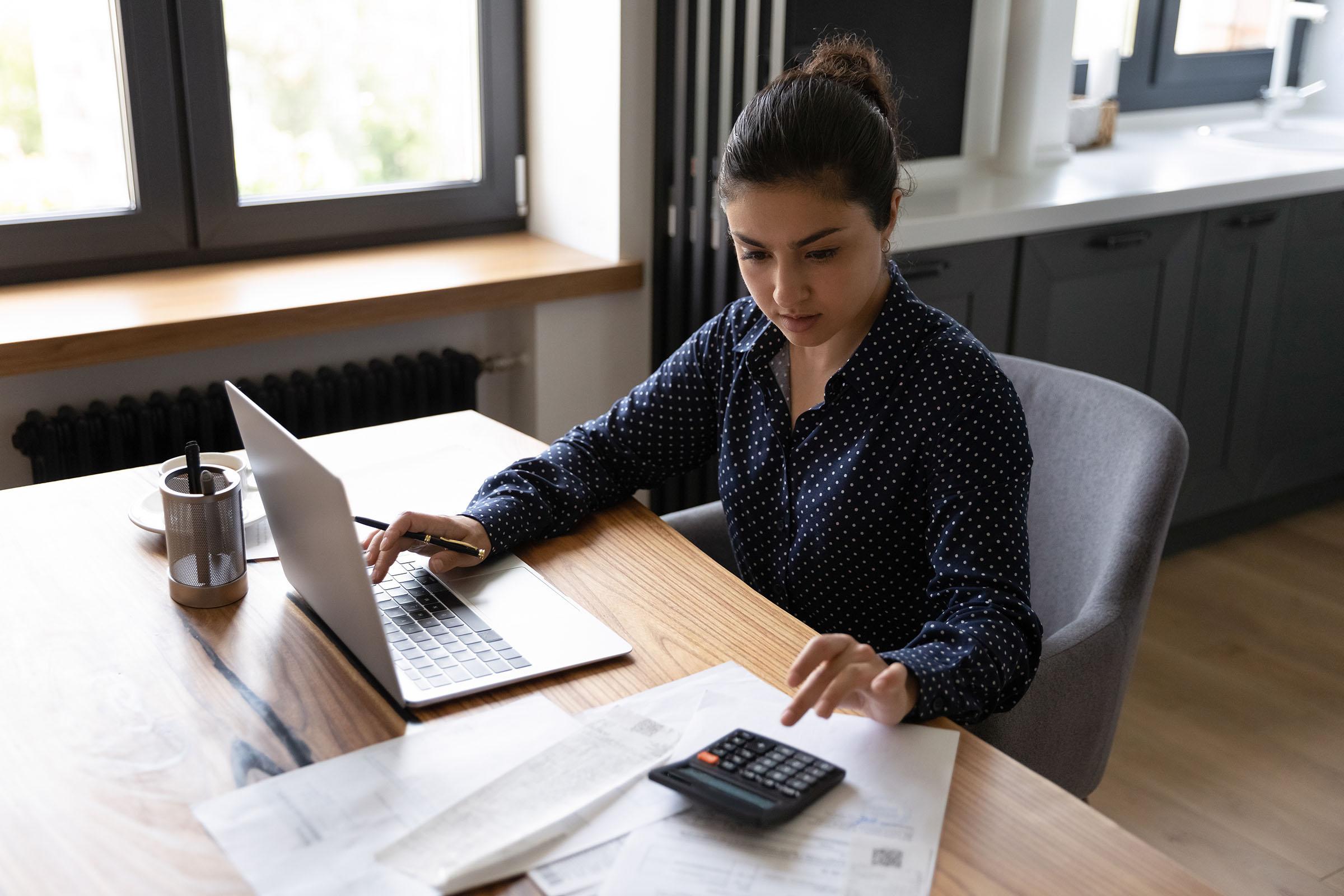 Woman in a dark polka dot blouse working at a table with a laptop, calculator, and financial documents in a modern kitchen.