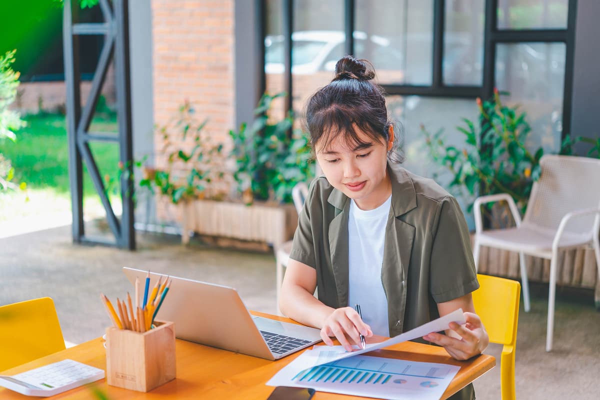 Woman working outdoors at a table with a laptop and papers, surrounded by greenery and a wooden pencil holder.