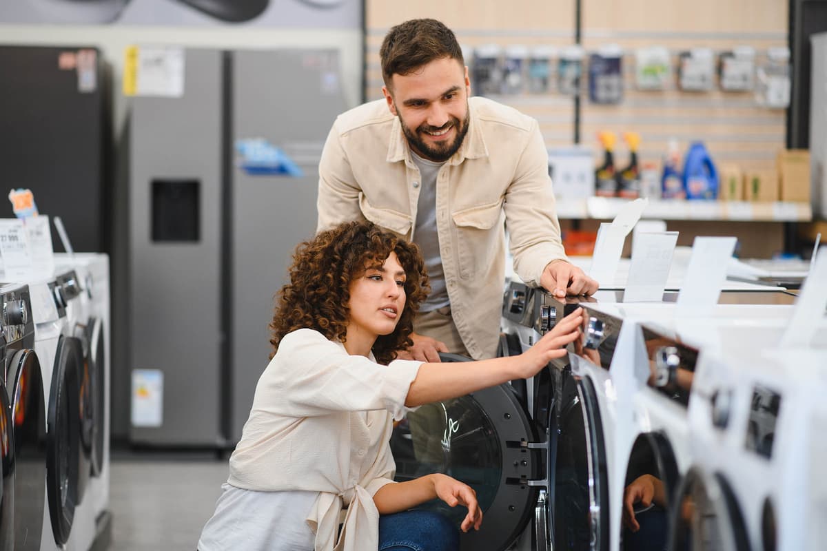 A couple examines washing machines in an appliance store, with the woman adjusting controls and the man overseeing.