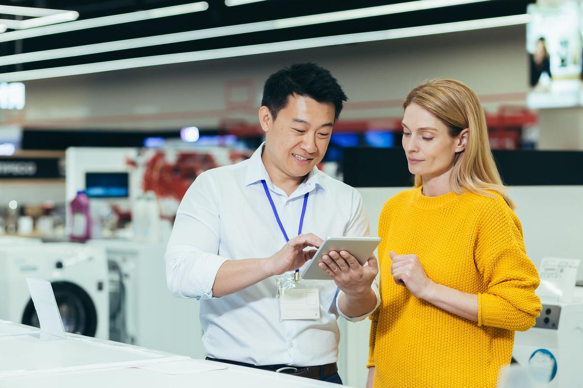 A male store employee shows a female customer information on a tablet in an appliance store, with washing machines in the background.