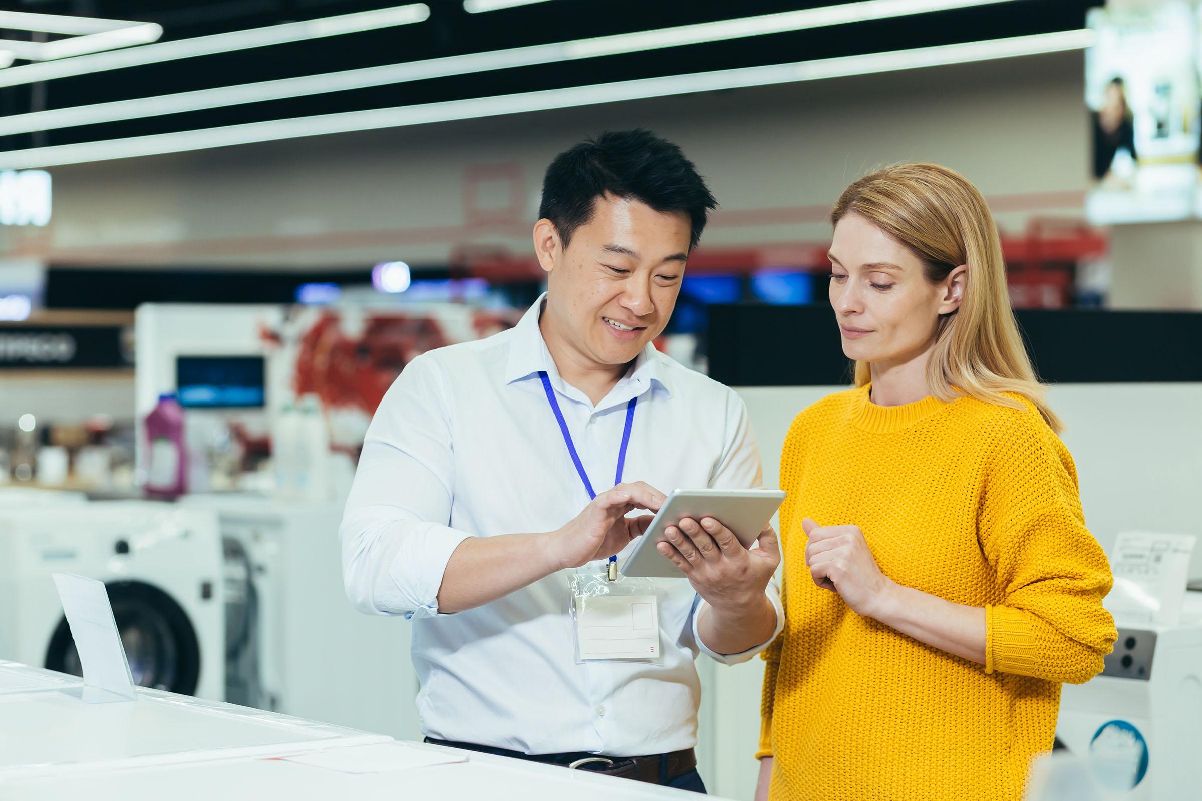 A male store employee shows a female customer information on a tablet in an appliance store, with washing machines in the background.