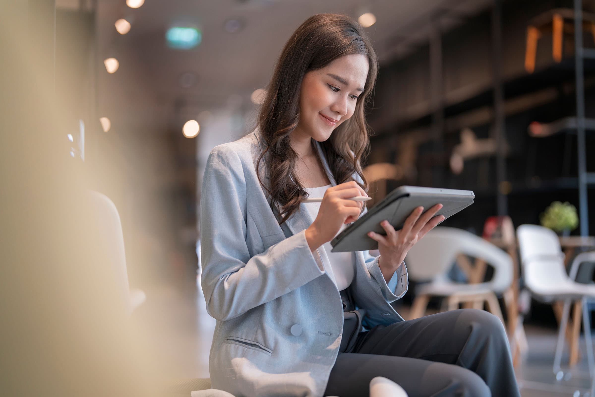 Woman in a blue blazer sits indoors, smiling while writing on a tablet. The background shows a modern, softly lit office environment.