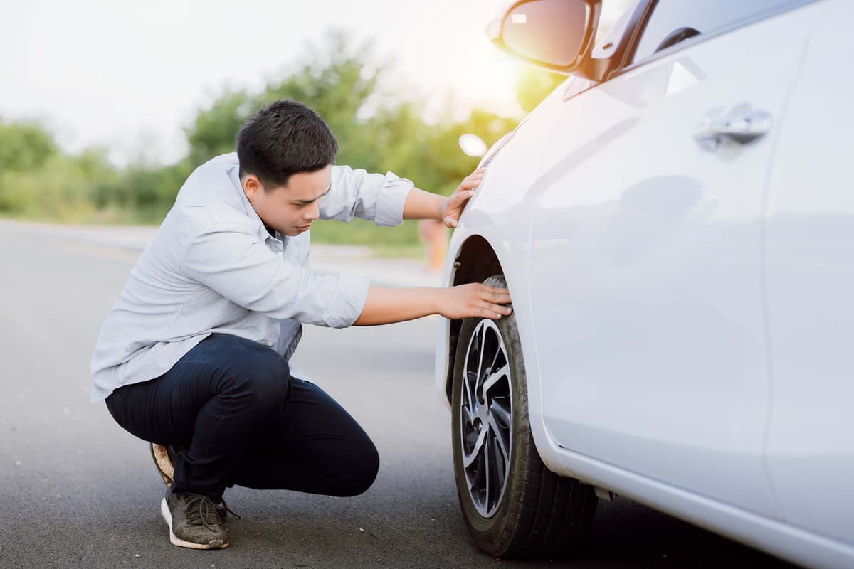 A person crouches down to inspect the tire of a white car parked on the road, with trees and sunlight in the background.