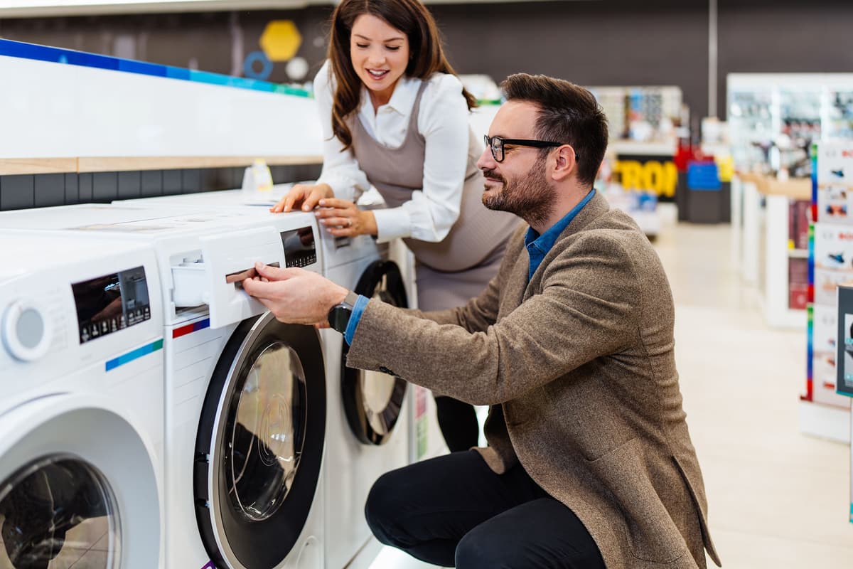 A man and woman in a store examine a washing machine, with the man pulling out the detergent drawer and the woman smiling beside him.