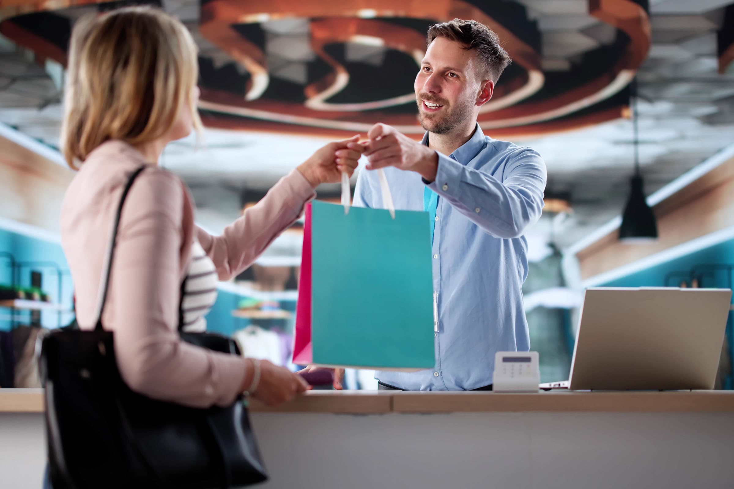 A cashier hands a shopping bag to a customer across the counter, both smiling. The scene is set in a modern, well-lit store lobby.