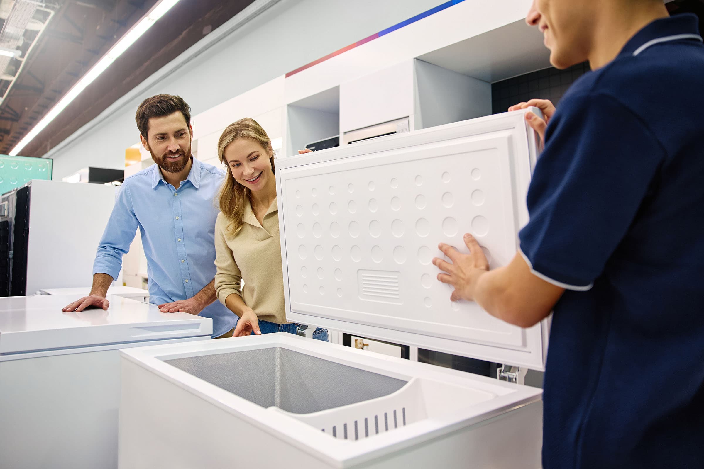 A couple examines a white chest freezer in a store as a sales associate opens the lid.