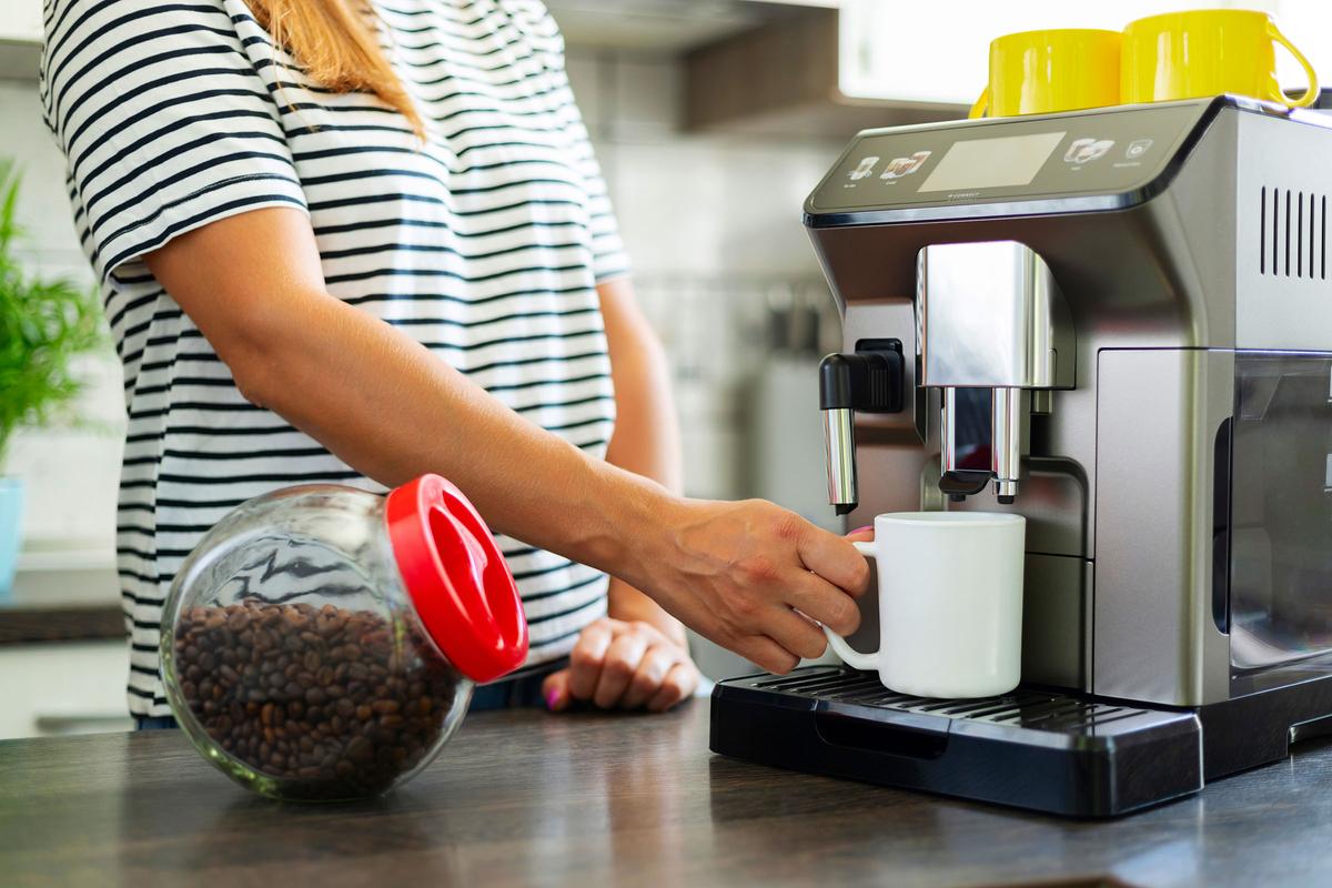 Person using a coffee machine to fill a white mug, with a jar of coffee beans and yellow mugs on the machine in a kitchen.
