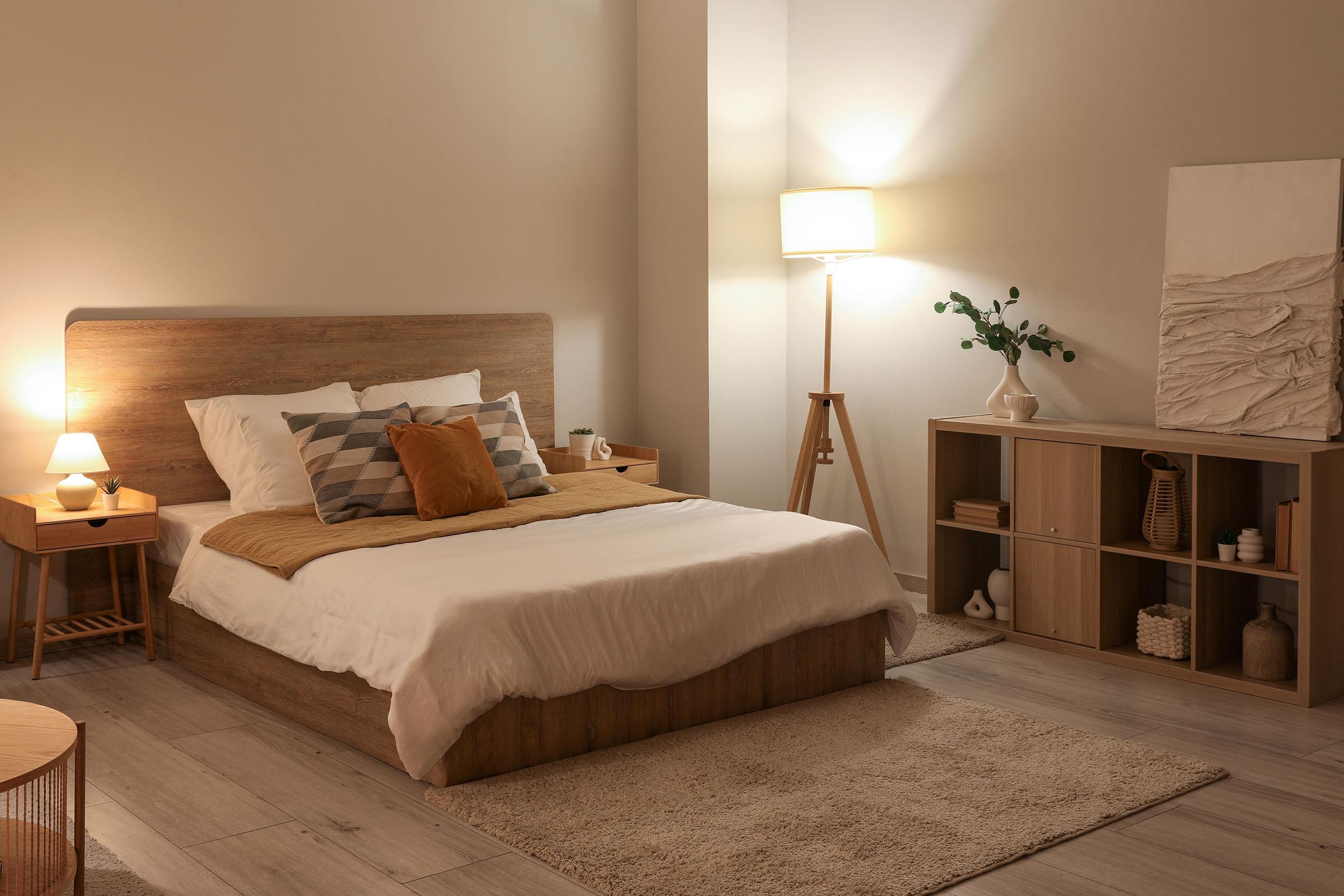 Cozy bedroom with a wooden bed frame, white and beige bedding, accent pillows, warm lighting, and minimalist decor.