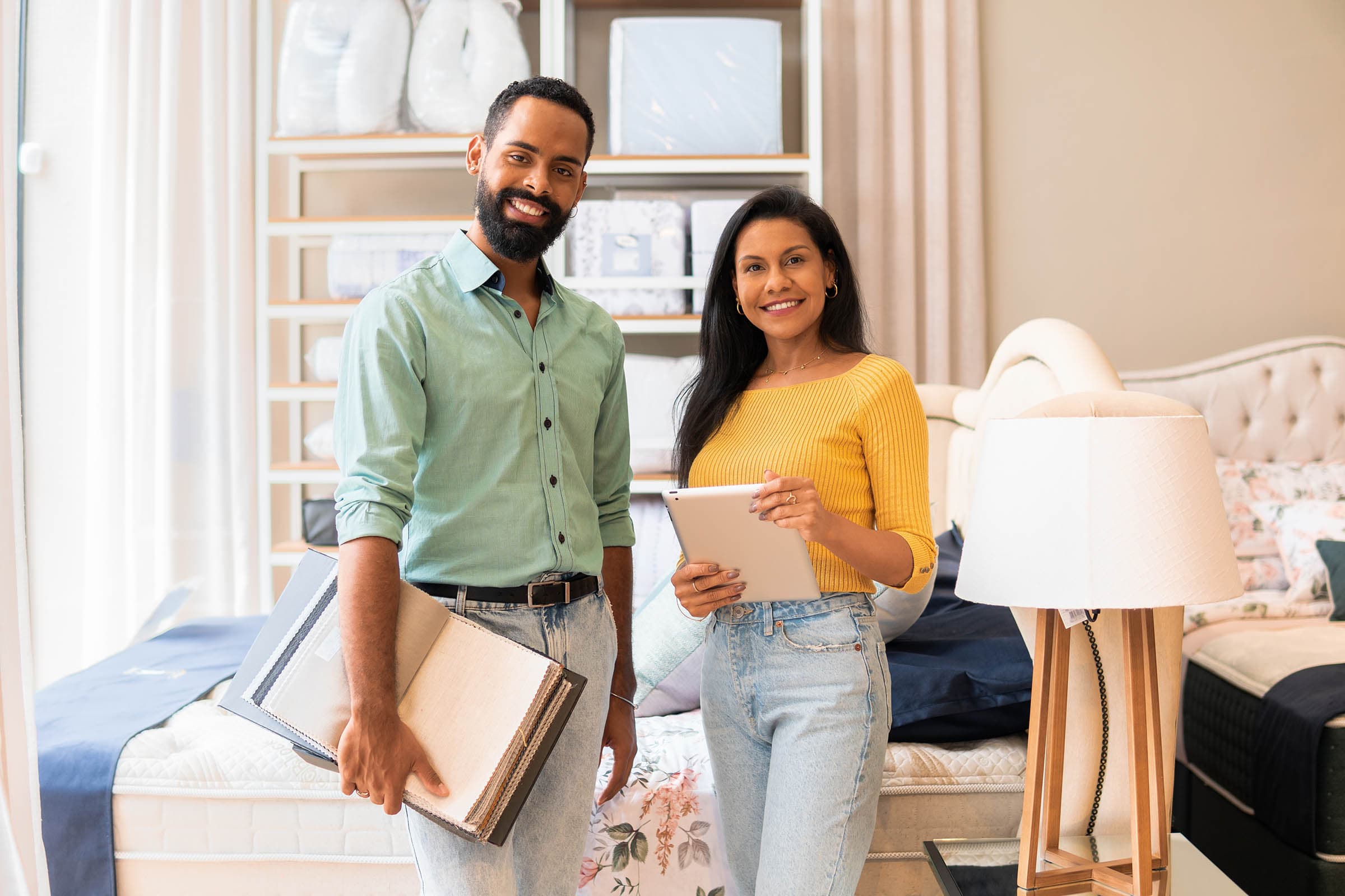A man and woman stand in a furniture store, holding a tablet and fabric samples, smiling. They are surrounded by bedding and a lamp.